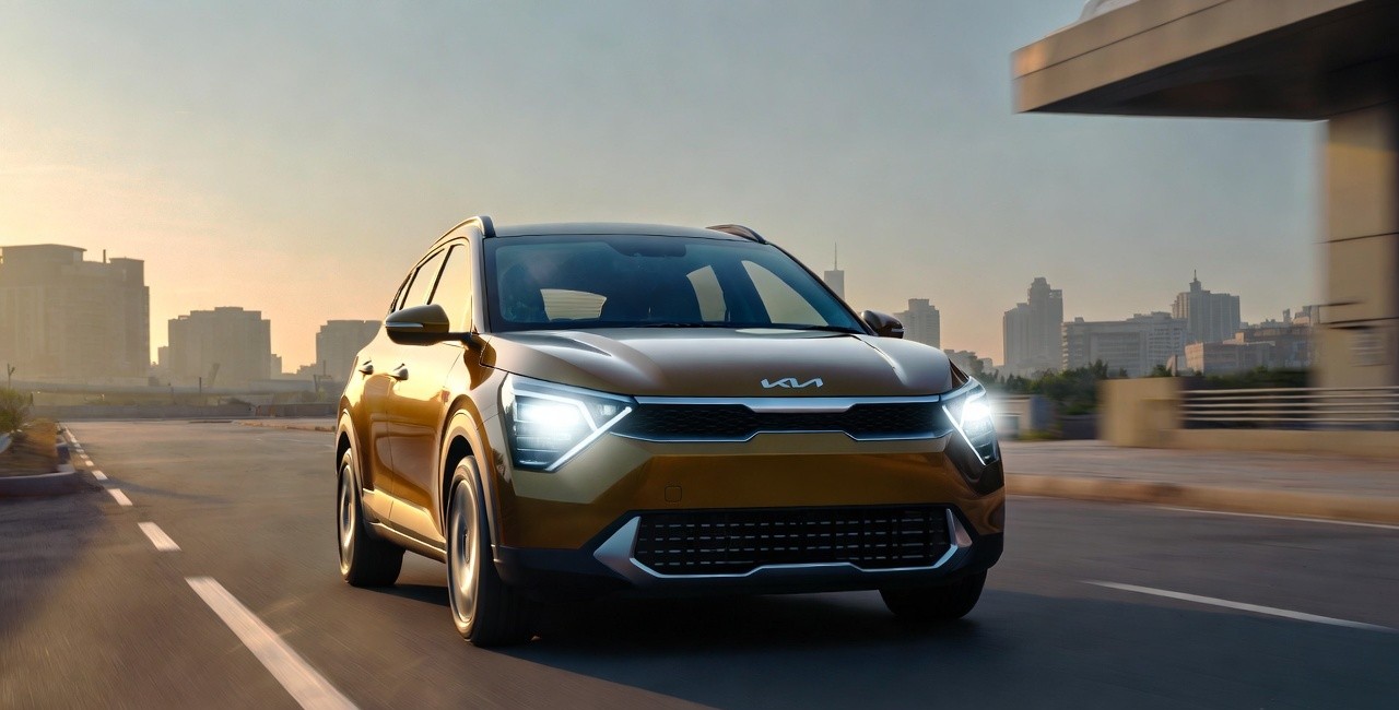 Kia Syros compact SUV parked in a modern Indian city at sunset with LED headlamps glowing.